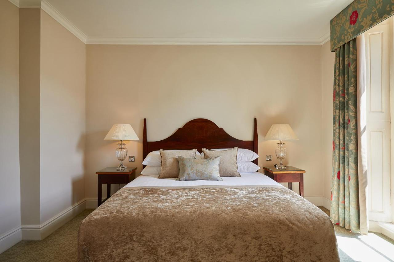 Macdonald Bath Spa Hotel Екстер'єр фото The photo shows a cozy hotel bedroom. It features a large bed with a plush, light-colored bedspread and several pillows. On either side of the bed, there are wooden nightstands with lamps providing soft lighting. The walls are painted a neutral color
