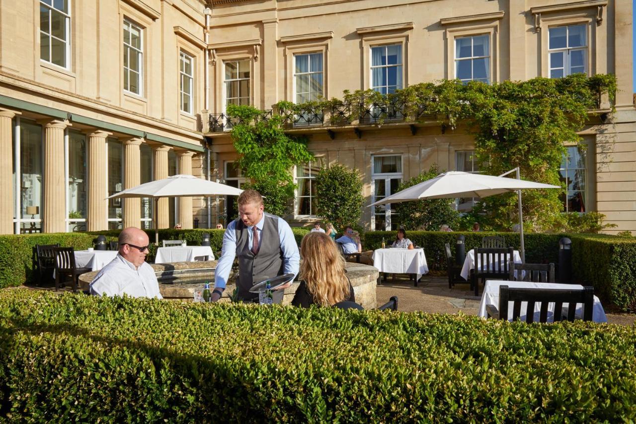 Macdonald Bath Spa Hotel Екстер'єр фото The photo depicts an outdoor dining area of a restaurant. In the foreground, there are neatly trimmed hedges surrounding the dining space. Two patrons are seated at a table; one is a man wearing sunglasses and the other is a woman facing him. A waite