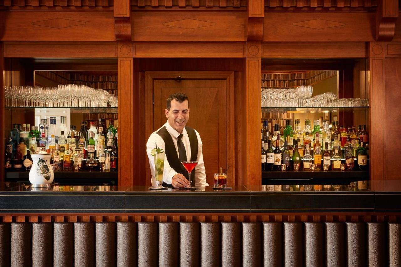 Macdonald Bath Spa Hotel Екстер'єр фото The photo depicts a bartender standing behind a polished bar. He is wearing a formal outfit with a vest and a bow tie, and he appears to be smiling as he presents a cocktail. Behind him is a well-stocked bar shelf filled with various bottles of liquo