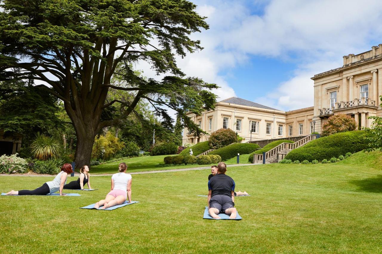 Macdonald Bath Spa Hotel Екстер'єр фото The photo shows a group of people engaging in a yoga session outdoors. They are positioned on yoga mats on a lush green lawn in front of a large, elegant building with columns. Behind them, there are trees and well-maintained gardens, contributing to