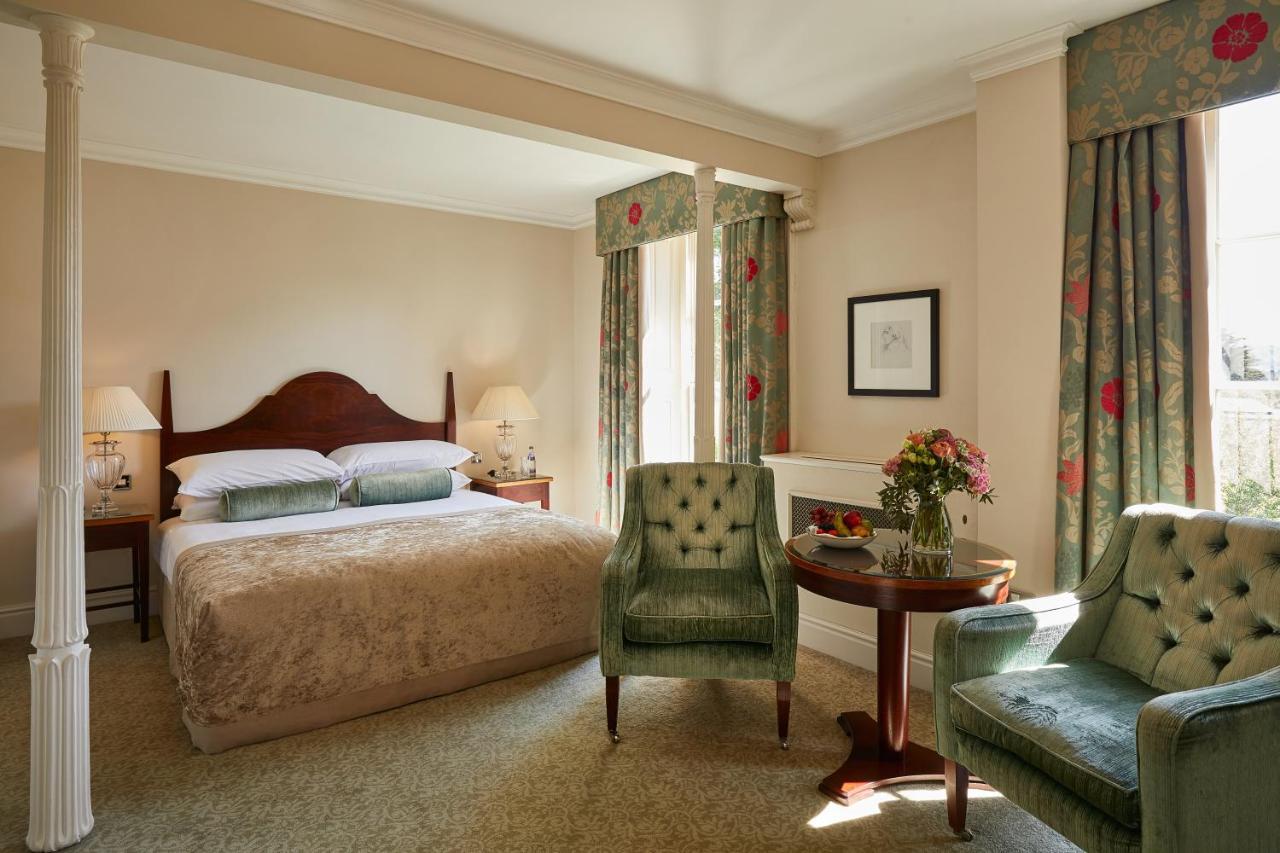 Macdonald Bath Spa Hotel Екстер'єр фото The photo shows a cozy hotel room featuring a large bed with a beige bedspread. There are two green armchairs positioned next to a small round table. The decor includes floral curtains and a neutral color palette on the walls. Soft lighting is provid