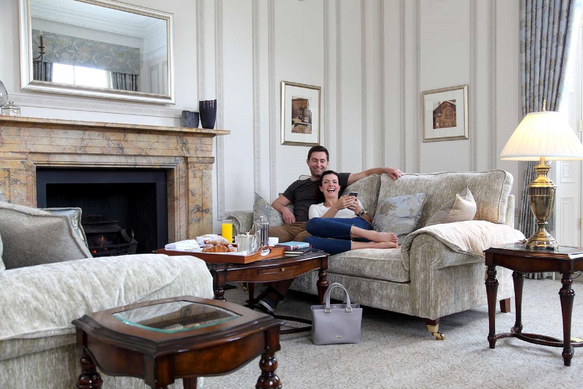 Macdonald Bath Spa Hotel Екстер'єр фото The photo shows a cozy living room setting where a couple is sitting together on a light-colored sofa. They appear relaxed, with the man sitting slightly behind the woman, who is smiling and looking at her phone. The room features elegant decor, incl