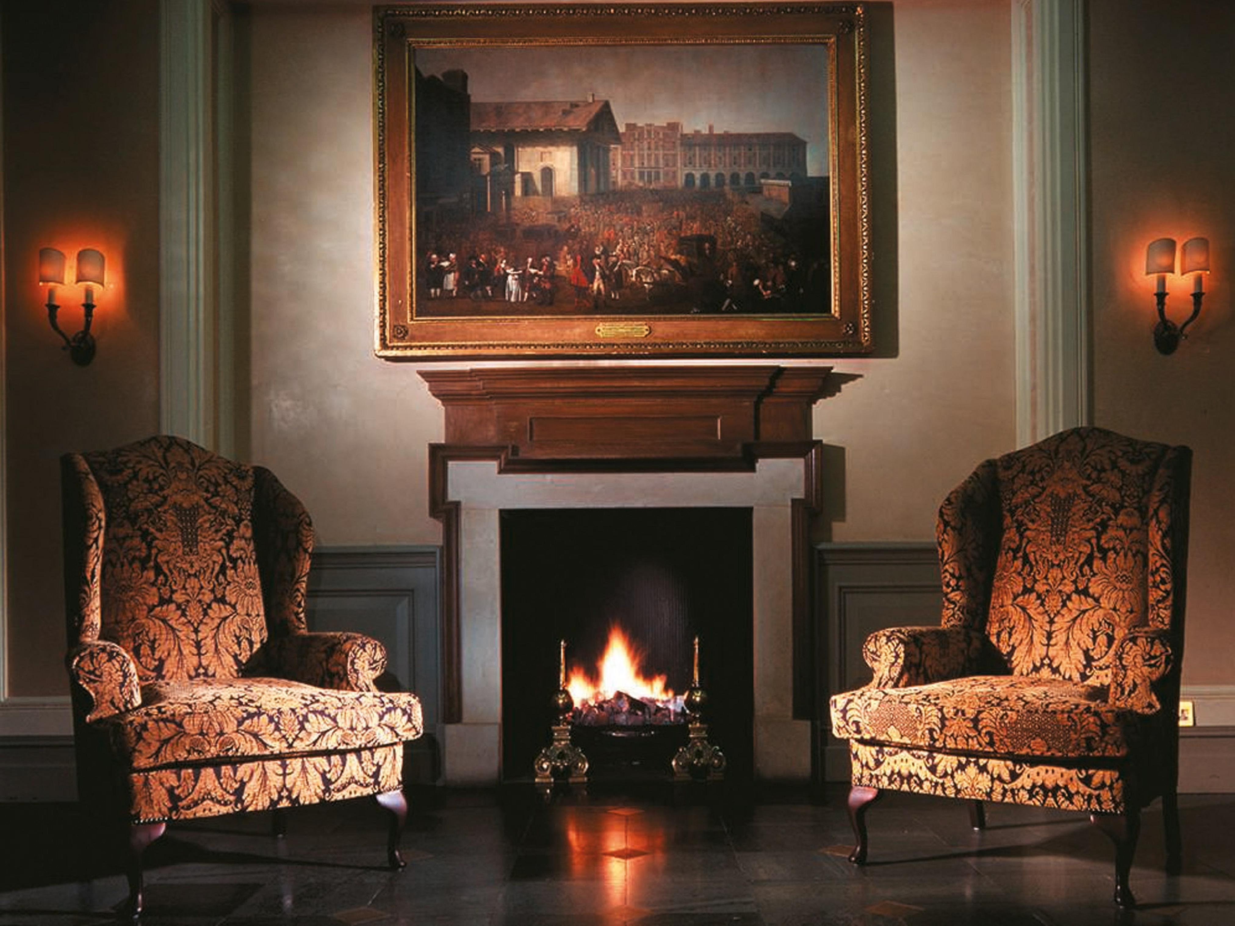 Macdonald Bath Spa Hotel Екстер'єр фото The image shows a cozy indoor setting featuring two ornate armchairs placed opposite a fireplace. Above the fireplace hangs a large painting in a decorative frame. The warm glow from the fire illuminates the room, and there are wall sconces providing