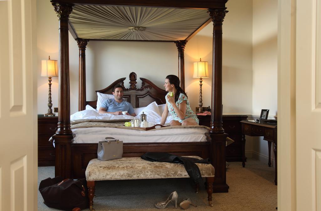 Macdonald Bath Spa Hotel Екстер'єр фото The image depicts a spacious, well-lit bedroom featuring a large four-poster bed with an ornate headboard. A man and a woman are sitting on the bed, appearing to engage in conversation. The woman is dressed in a patterned outfit, while the man is wea